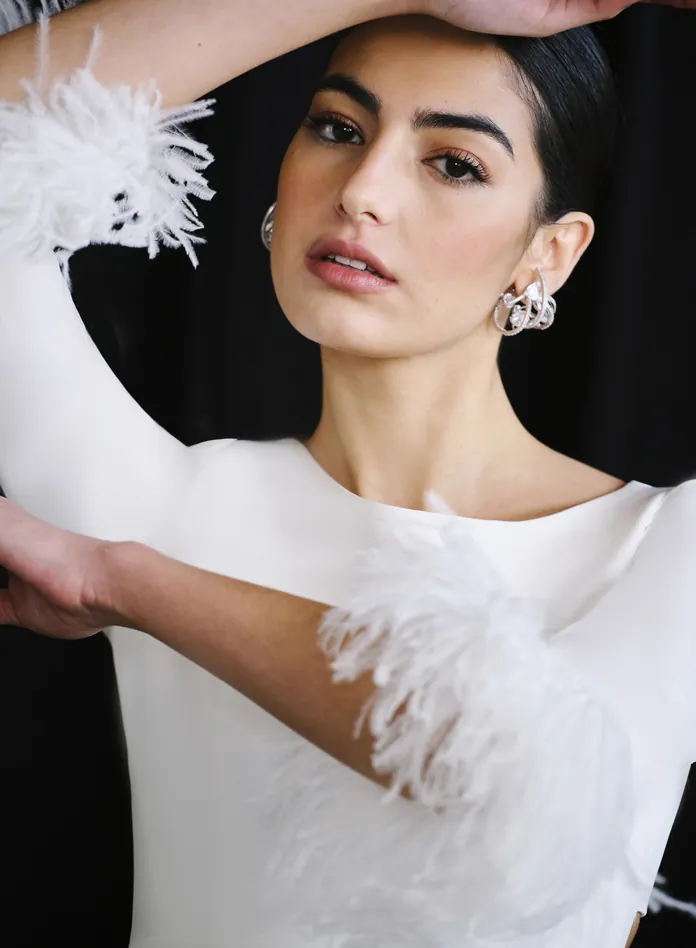 Vestido de novia confeccionado en crepe, con líneas limpias que abrazan la silueta con elegancia y sobriedad. Las plumas en los puños aportan un toque sofisticado y glamuroso, perfecto para una novia con personalidad. La abertura en la falda y el original escote en la espalda aportan sensualidad y hacen que este diseño no pase desapercibido.