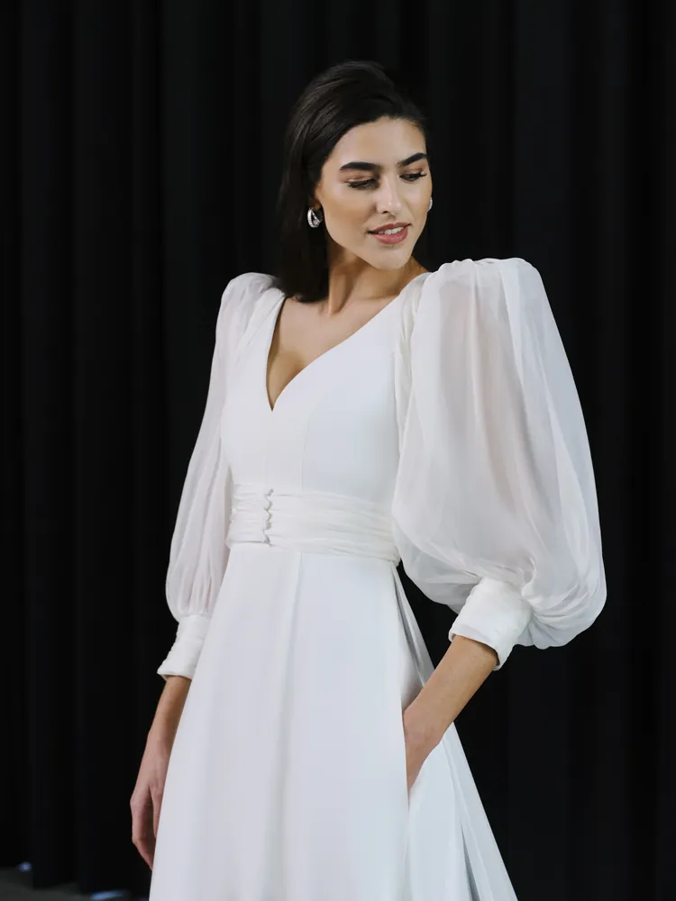 Vestido de novia versátil que combina dos looks en uno. Confeccionado en crepe y de cuerpo entallado, presenta un escote en pico y espalda abierta. La falda destaca por su caída fluida y añade un toque divertido con los bolsillos. El vestido se complementa con un abrigo desmontable de organza, caracterizado por unas mangas abullonadas acabadas con adorno de botones en los puños.