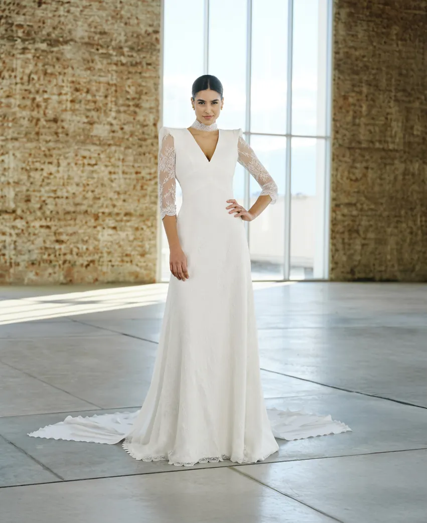 Vestido de novia que une elegancia y distinción en un diseño majestuoso confeccionado en crepe y chantilly. Su silueta femenina se adorna con un sofisticado fajín triangular que enmarca la cintura, mientras que el delicado encaje chantilly cubre el cuerpo, las mangas y la falda, aportando un aire romántico. El detalle más icónico es su choker bordado en guipur, unido a unas caídas de gasa desmontables que completan el look con una caída única. Cleopatra es la elección perfecta para novias que buscan impactar con seguridad y una esencia atemporal. Reservar cita