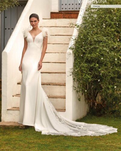 Vestido de novia de corte sirena y estilo romántico. confeccionado en crepe y combinado con pedrería y aplicaciones florales en 3D. Presenta un sensual escote en V. hombros adornados con plumas y una espalda ilusión en tul adornada con aplicaciones. dejando paso a una falda muy femenina con una espectacular cola muy trabajada que combina pedrería con flores bordadas 3D. Todo un diseño de costura muy glamuroso y sexy.