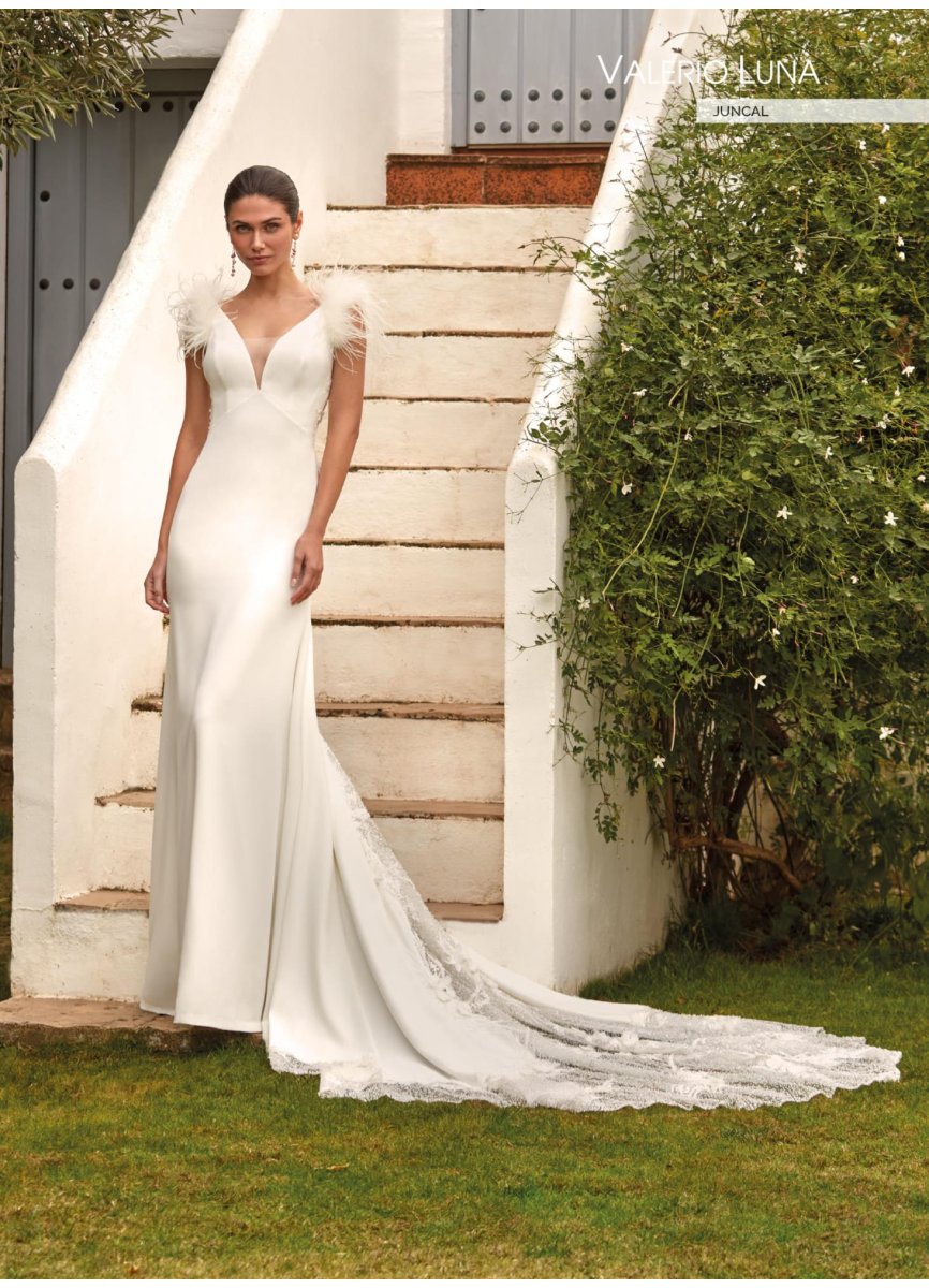 Vestido de novia de corte sirena y estilo romántico. confeccionado en crepe y combinado con pedrería y aplicaciones florales en 3D. Presenta un sensual escote en V. hombros adornados con plumas y una espalda ilusión en tul adornada con aplicaciones. dejando paso a una falda muy femenina con una espectacular cola muy trabajada que combina pedrería con flores bordadas 3D. Todo un diseño de costura muy glamuroso y sexy.