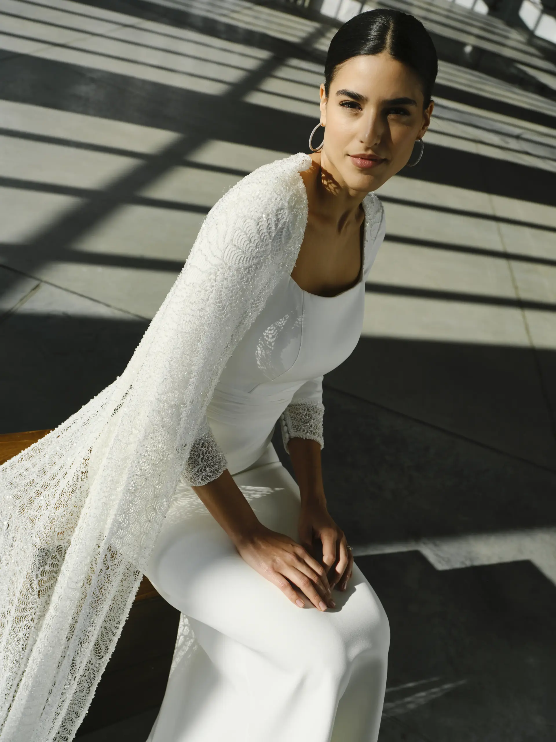 Vestido de novia confeccionado en crepe que presenta un escote cuadrado. Tanto sus hombros como sus puños están realizados en tul bordado con pedrería. Un delicado fajín de gasa, marca la cintura y la espalda sorprende con cut out y falda con botones joya. La capa también en pedrería que nace desde los hombros, te hará brillar como nunca.