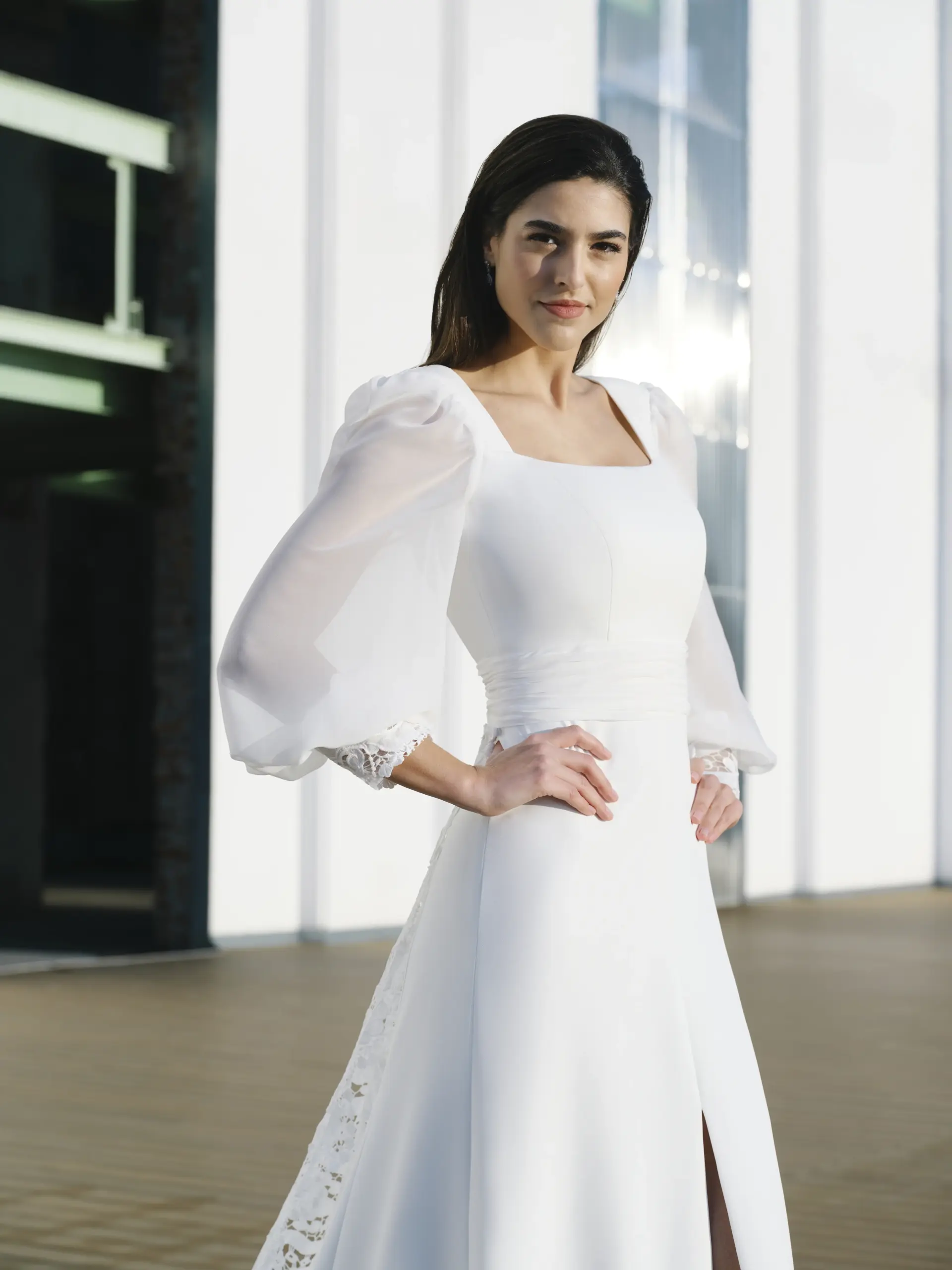 Vestido de novia en crepe con escote cuadrado, mangas abullonadas en gasa y espalda abierta en forma de V. Fajín fruncido en gasa y sobrecola troquelada que aporta textura y un efecto visual único.