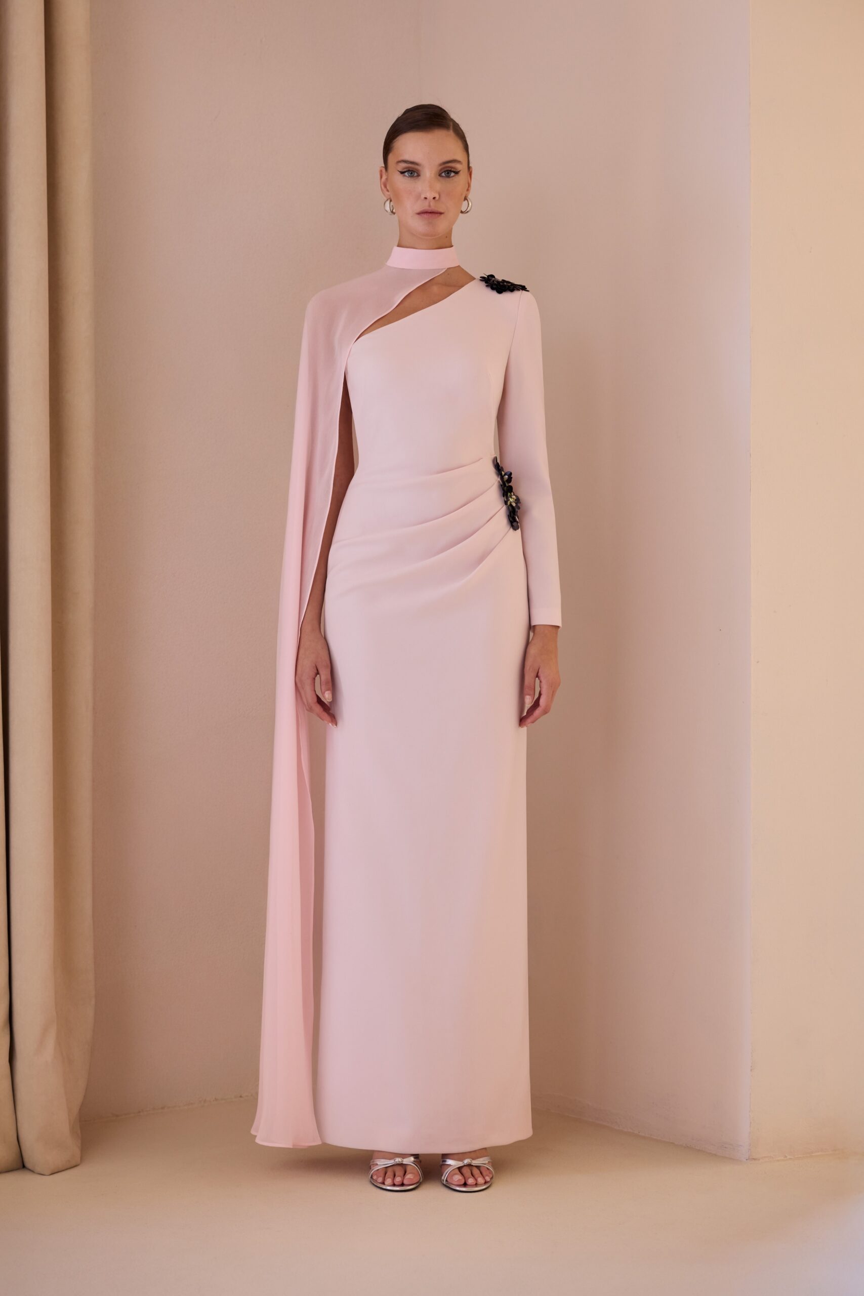 Vestido largo de corte recto y silueta estilizada que destaca por su elegancia atemporal. Presenta original escote asimétrico con cuello alto, manga larga y una delicada capa lateral que aporta movimiento y sofisticación. El detalle joya en el hombro añade un toque exclusivo. Ideal para eventos, bodas, graduaciones o celebraciones especiales donde quieras brillar con clase.