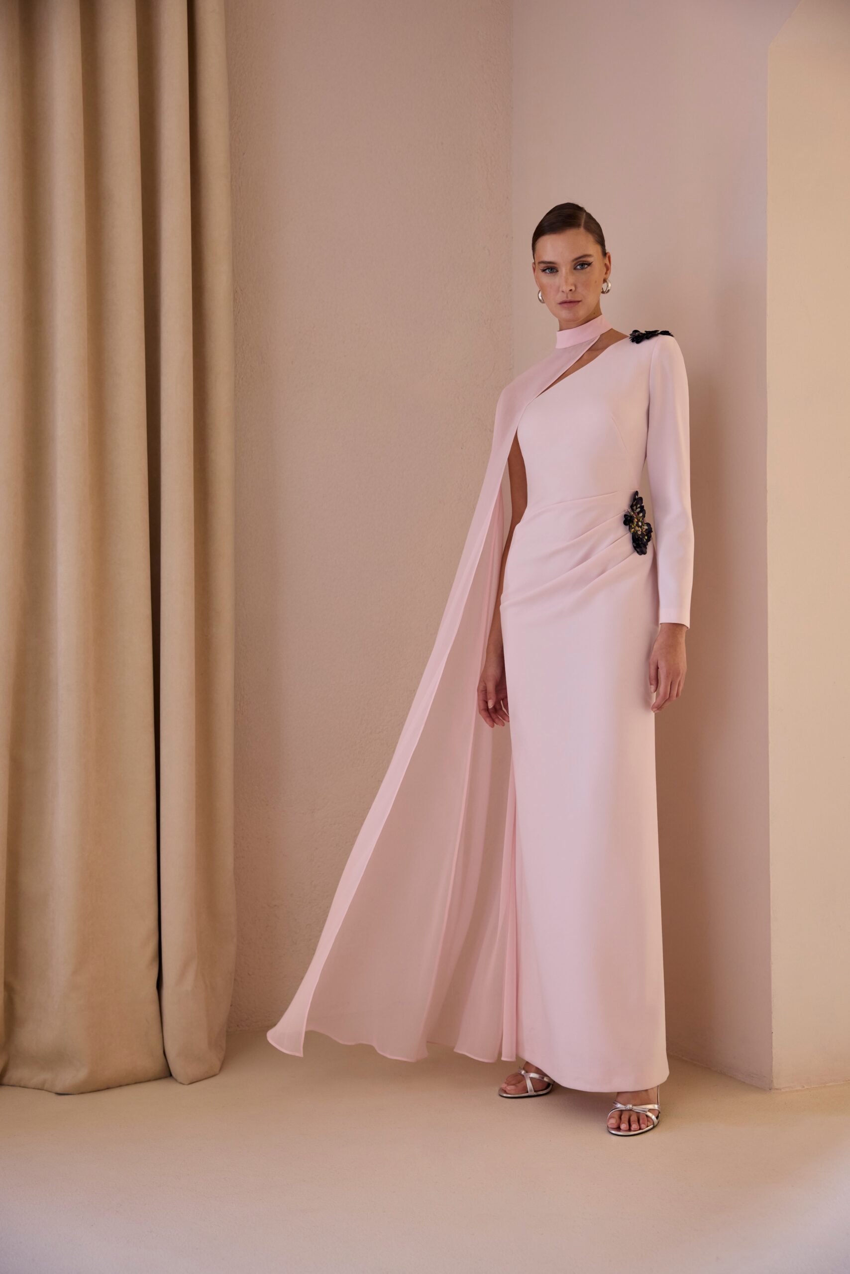 Vestido largo de corte recto y silueta estilizada que destaca por su elegancia atemporal. Presenta original escote asimétrico con cuello alto, manga larga y una delicada capa lateral que aporta movimiento y sofisticación. El detalle joya en el hombro añade un toque exclusivo. Ideal para eventos, bodas, graduaciones o celebraciones especiales donde quieras brillar con clase.